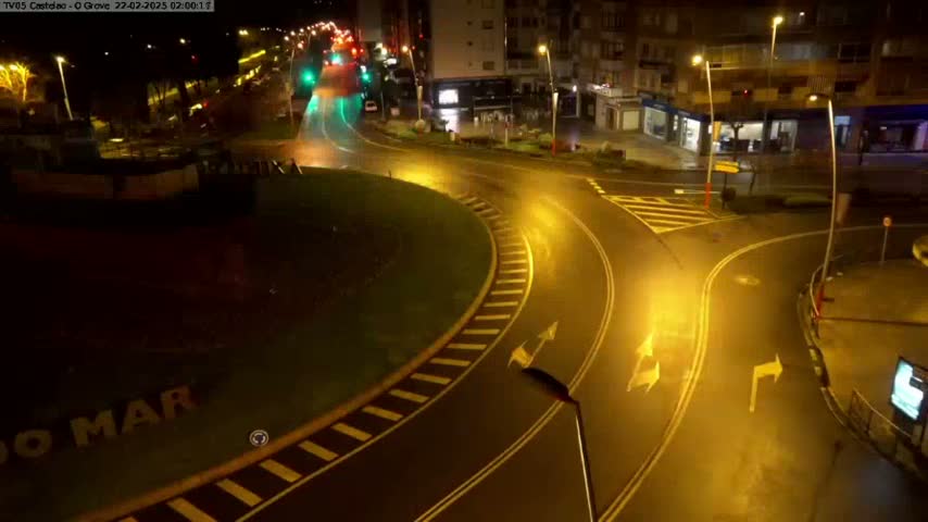 A wet roundabout at night, illuminated by streetlights, with vehicles moving through it and buildings visible in the background.
