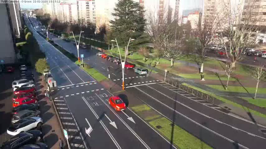 A sunny day reveals a multi-lane road with cars driving in both directions, bordered by parked cars and a grassy area with trees and buildings in the background.