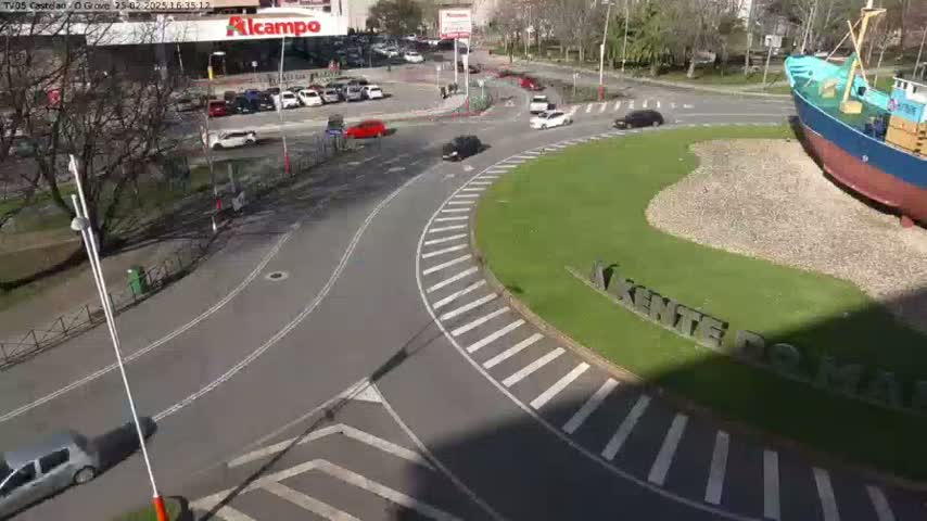 A sunny day reveals a roundabout with several cars, a large boat sculpture, and a grassy area, all surrounded by buildings and trees.