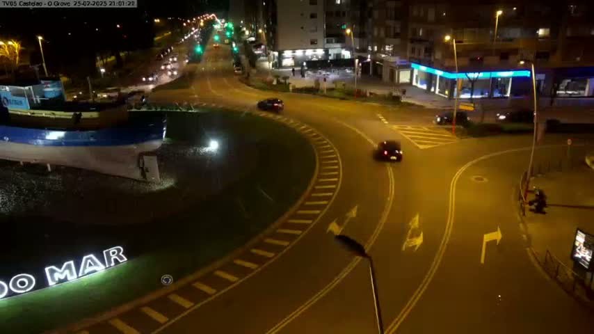 A nighttime view of a roundabout with a large boat sculpture at its center, several cars driving around it, and surrounding buildings illuminated.