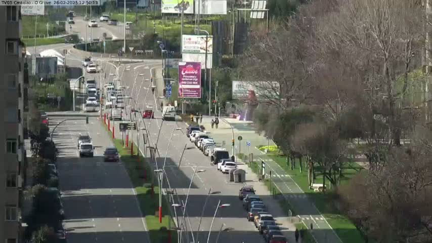 A sunny day shows a multi-lane road with cars driving in both directions, lined with bare trees and a bike path.
