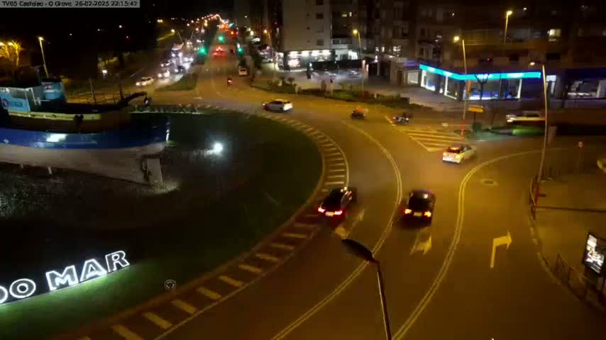 A nighttime aerial view shows a roundabout with several cars driving around it, near a large model of a boat, and next to buildings.