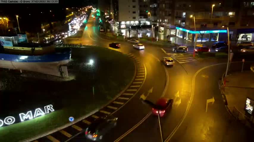 A wet, nighttime roundabout scene shows several cars navigating the circular roadway, with a large boat-shaped structure and buildings visible in the background.