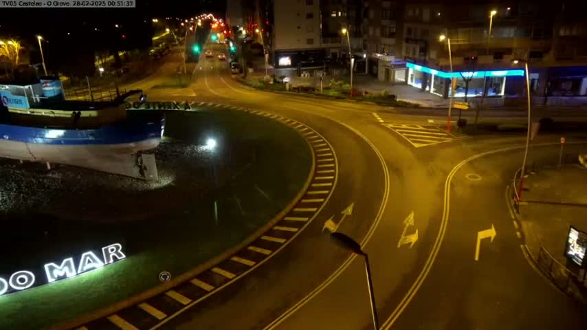 At night, a roundabout with a large boat sculpture at its center is illuminated by streetlights, with a few cars visible on the roads leading to it.