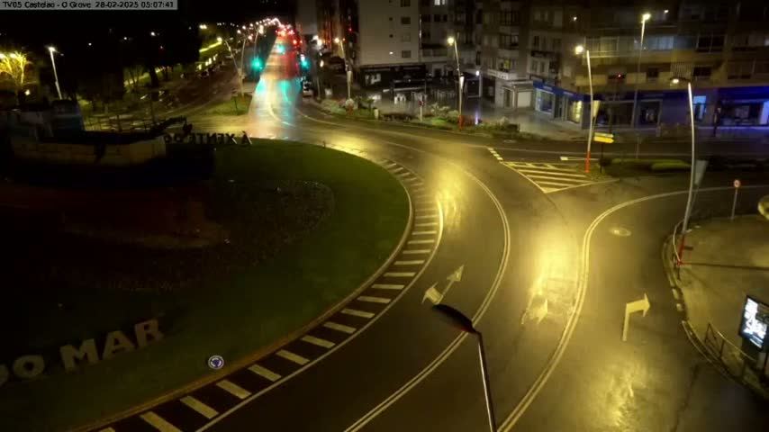 A wet roundabout at night, illuminated by streetlights, with a few vehicles visible in the distance and a boat-like structure in the foreground.