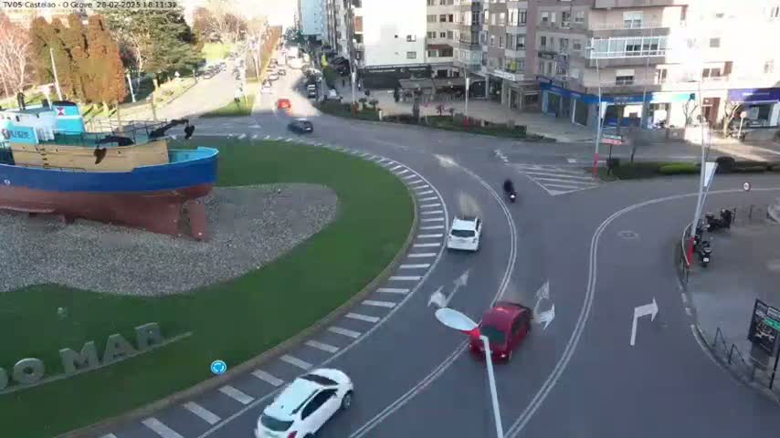A large model of a fishing boat sits in a roundabout with several cars and motorcycles navigating the curved roadways on a sunny day.
