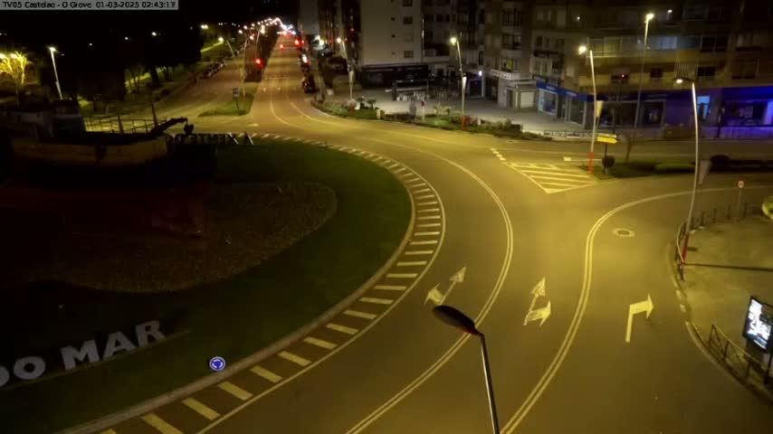 An empty roundabout at night, illuminated by streetlights, with a boat visible in the background.
