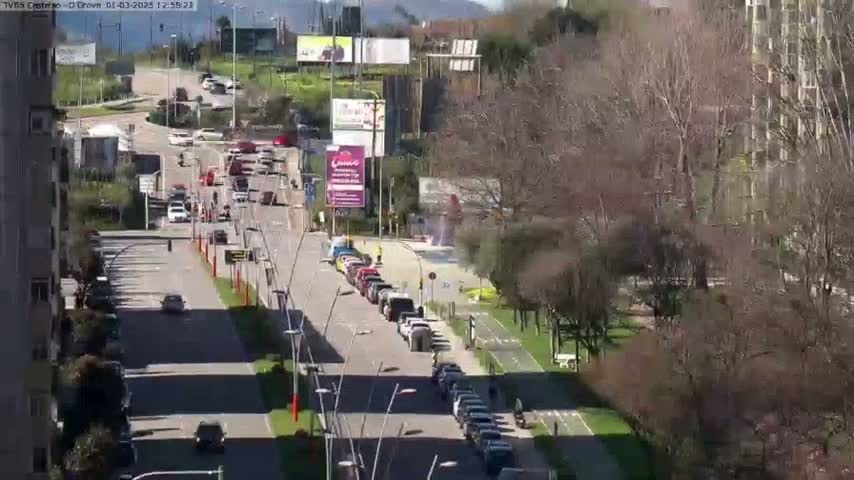A sunny day shows a multi-lane road with cars driving in both directions, lined with buildings and trees, and a bike path adjacent to the road.