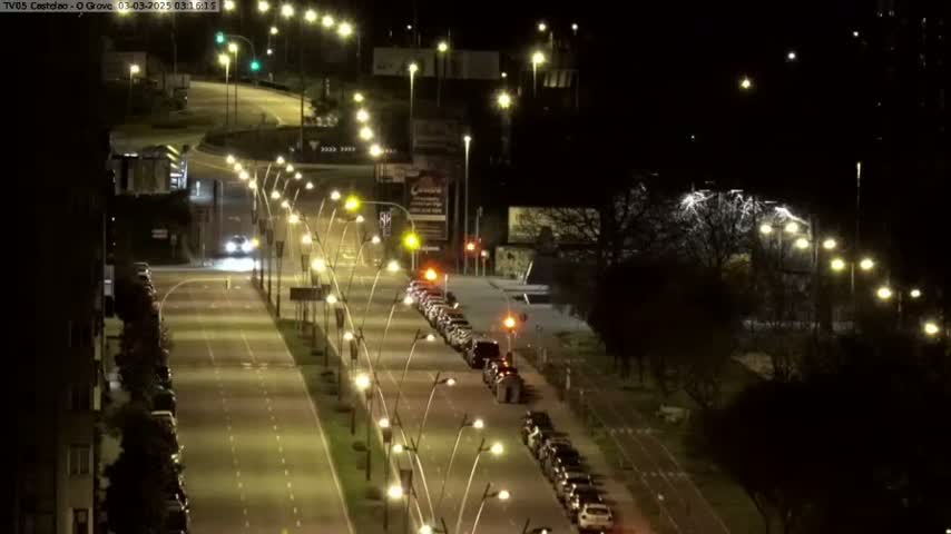 A nighttime, well-lit city street scene shows cars driving on a multi-lane road flanked by parked cars and streetlights, with trees lining the sides of the road.