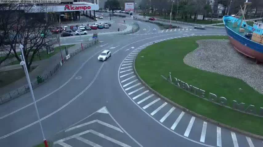 A white car drives through a roundabout in a city on an overcast day, near a large ship statue.