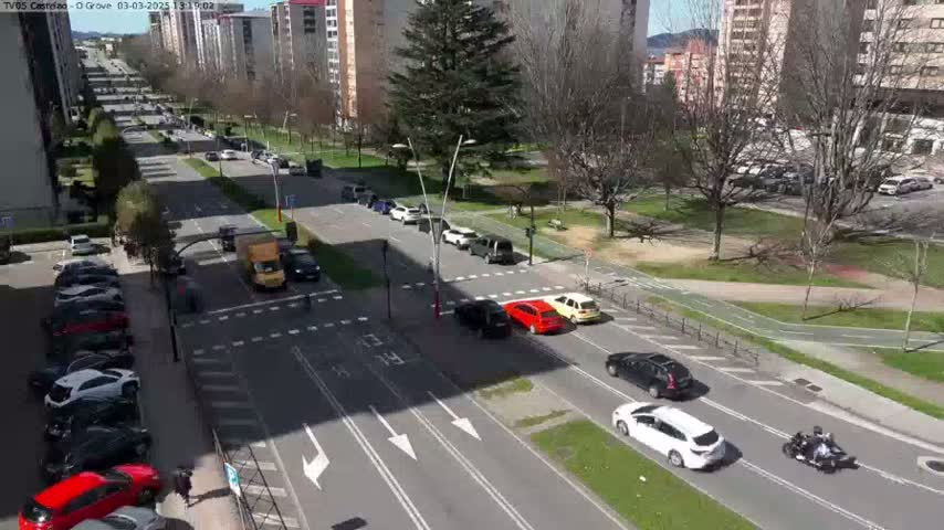 A sunny day shows a high-angle view of a city intersection with multiple cars driving on a multi-lane road, with parked cars lining a side street, and a few pedestrians walking nearby.