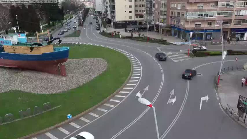 A large model of a fishing boat sits in a grassy roundabout at a roadway intersection, with several cars navigating the area on a partly cloudy day.