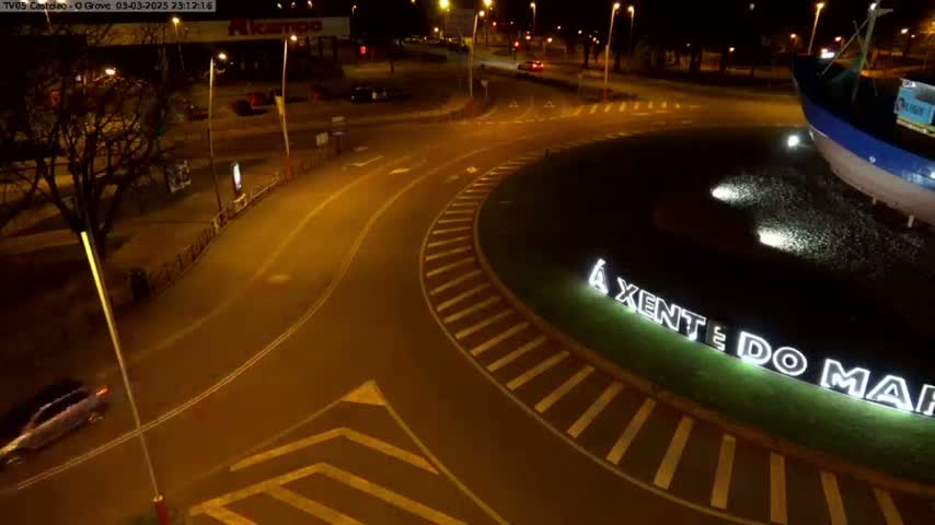 A nighttime aerial view shows a roundabout with a partially visible boat sculpture and illuminated lettering, and a car driving on the road.