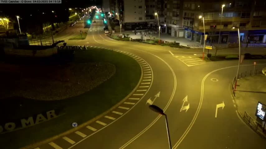 An empty roundabout and roads at night, illuminated by streetlights, with a boat visible in the background.
