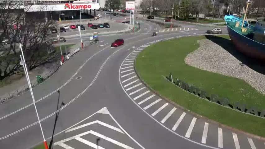 A sunny day shows a curved roadway with crosswalks, a few cars, and a large ship partially visible at the bottom right, all surrounded by green grass and trees.