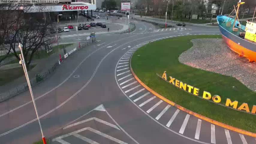 An aerial view shows a roundabout with a large ship sculpture in the center, surrounded by roads and parking areas, under a partly cloudy sky.