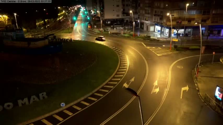 A wet, nighttime roundabout is illuminated by streetlights, with a few cars visible on the roadway and buildings in the background.