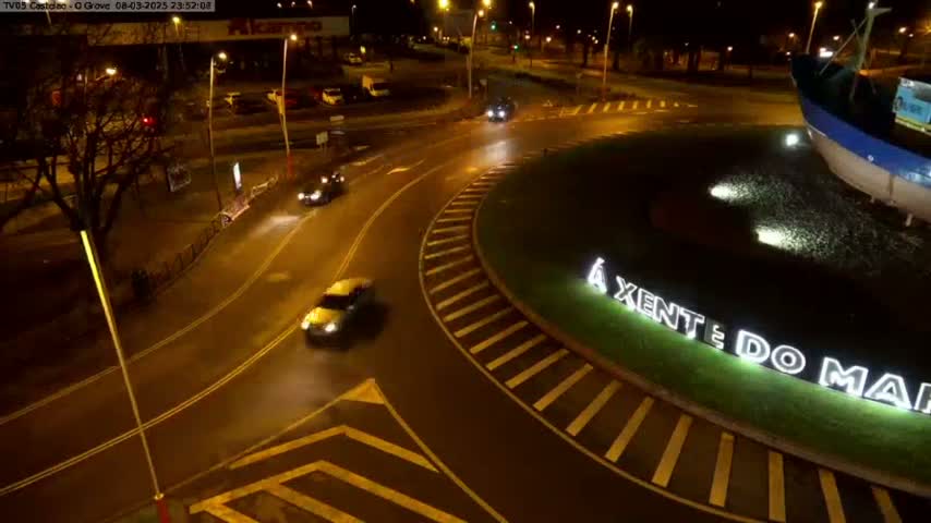 At night, several cars are driving around a roundabout with a large illuminated sign and a model of a fishing boat in the center, under wet conditions.