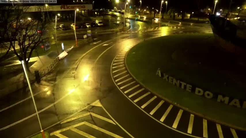 A wet, nighttime scene shows a roundabout with cars parked nearby and streetlights illuminating the rain-slicked asphalt.