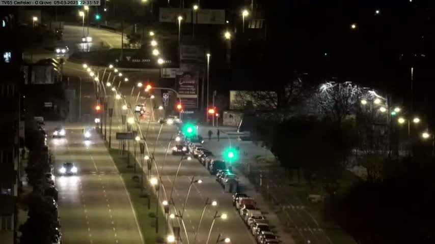 A nighttime, brightly lit road with multiple lanes of slow-moving traffic, bordered by trees and buildings.