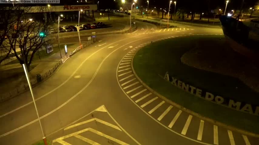 An empty roundabout at night, illuminated by streetlights, is surrounded by a parking lot and trees.