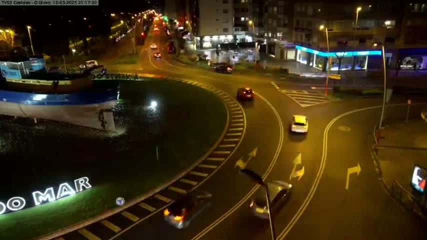 A nighttime aerial view of a roundabout with several cars driving around it, near a body of water containing a boat.