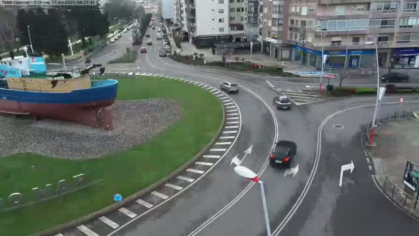 A large, blue and brown ship sits on the grass next to a roundabout where several cars are driving on a wet, gray day.