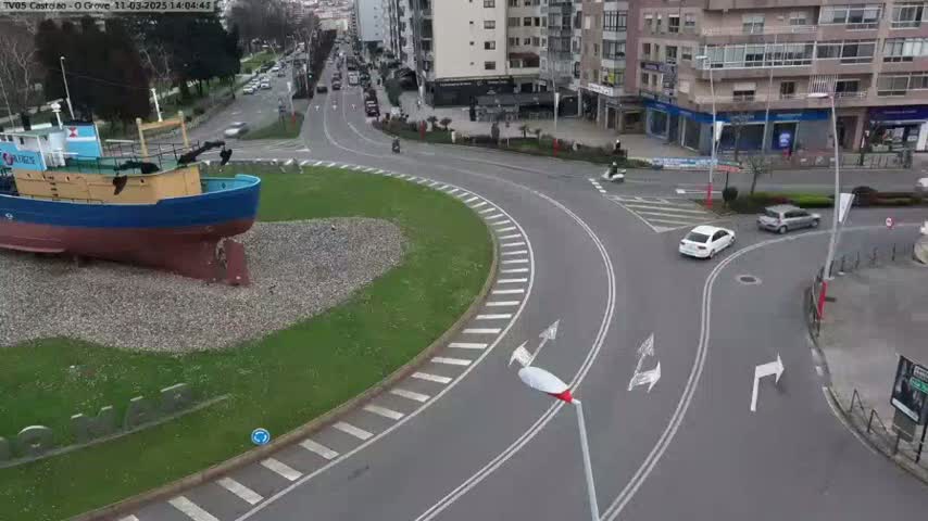 A large model of a fishing boat sits in a grassy roundabout, surrounded by a paved roadway with several cars and motorcyclists navigating the curves on a partly cloudy day.