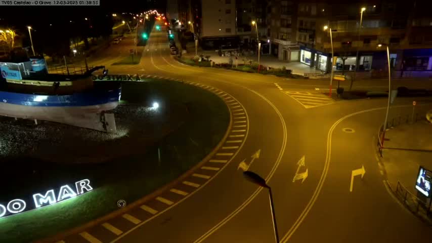 A nighttime aerial view shows a roundabout with a boat statue at its center, surrounded by illuminated streets and buildings.