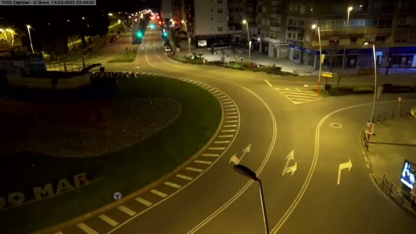 A nighttime aerial view shows a mostly empty roundabout and intersecting roads with buildings in the background, illuminated by streetlights.
