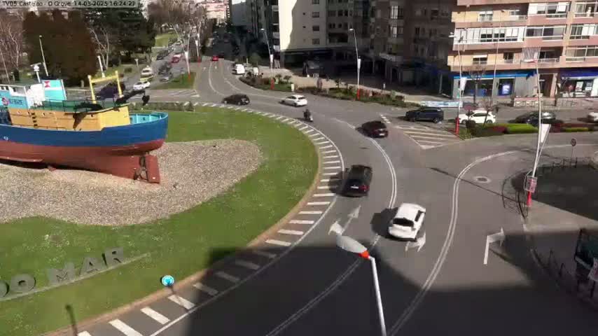 A model of a blue and yellow fishing boat sits in a grassy roundabout, with several cars navigating the sunny streets around it.