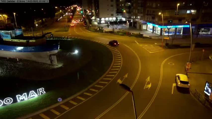 At night, a roundabout with a few cars and a large sculpture of a boat is visible, illuminated by streetlights.