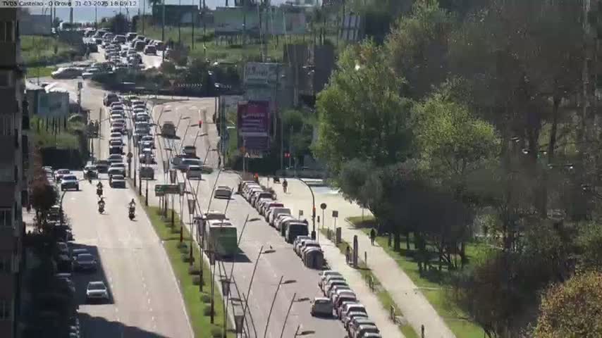 A sunny day reveals a multi-lane road with heavy traffic, including cars and motorcycles, alongside a tree-lined path.