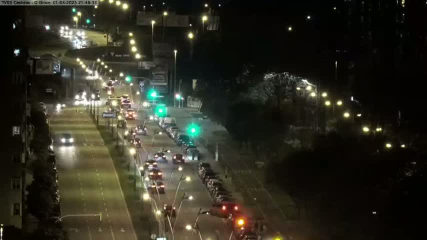 A nighttime, high-angle view shows a multi-lane road with moderate traffic flowing in both directions, lined with streetlights and trees.