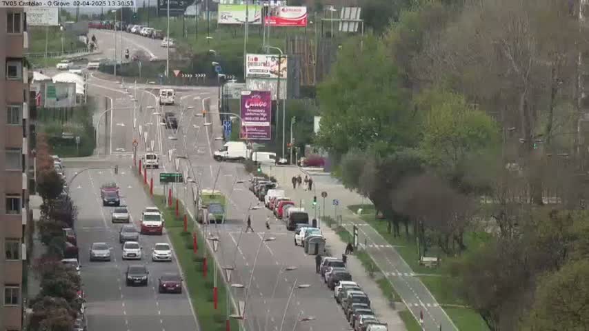A partly cloudy day reveals a multi-lane road with several cars driving in both directions, flanked by buildings and greenery, including a bike path.