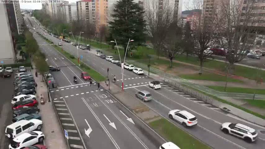 A city street intersection with several cars and pedestrians, under a partly cloudy sky.