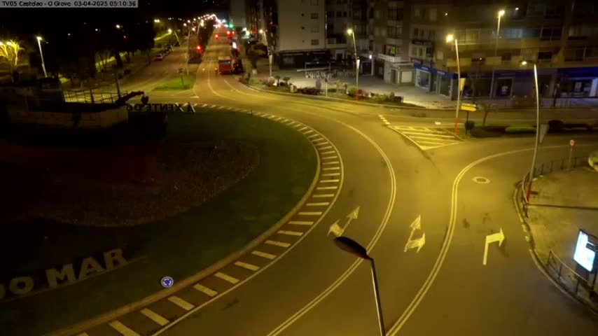 A nighttime aerial view shows a mostly empty roundabout and intersecting roads, illuminated by streetlights, with a boat visible in the lower left corner.