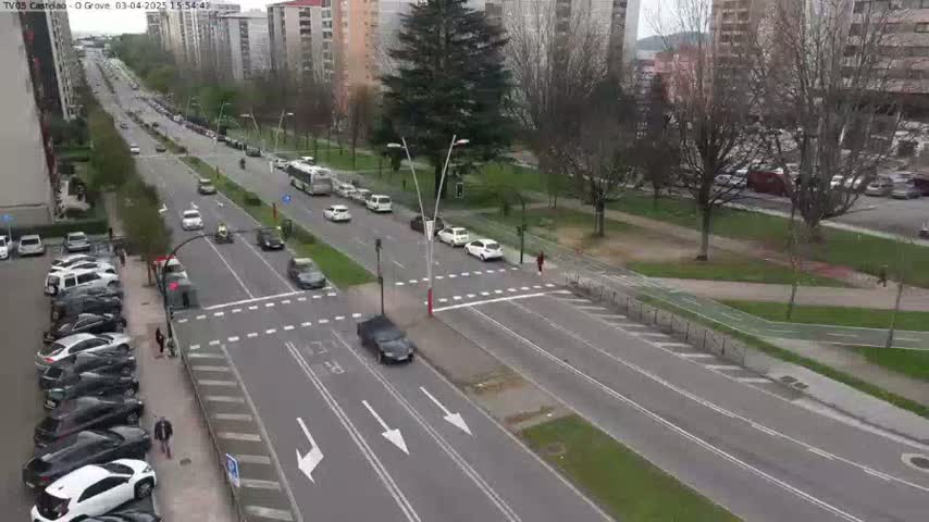 A multi-lane road with cars driving in both directions, parked cars lining the side of the road, and buildings and trees in the background on a partly cloudy day.