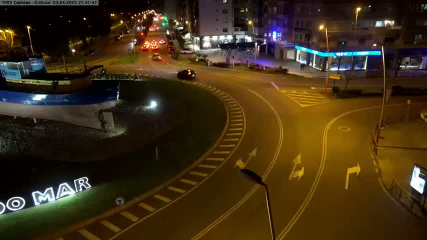 A nighttime aerial view shows a roundabout with a few cars, a large boat sculpture, and buildings in the background under illuminated streetlights.