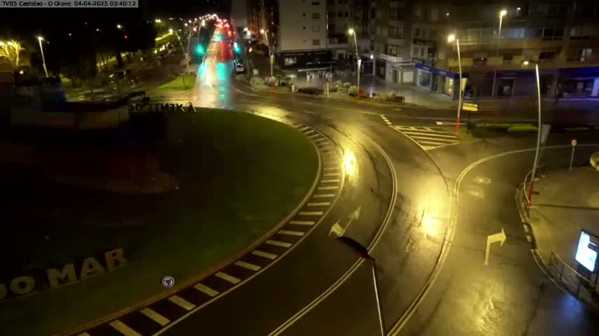 A wet, nighttime view of a roundabout in a town, with several vehicles visible on the roads and buildings in the background.