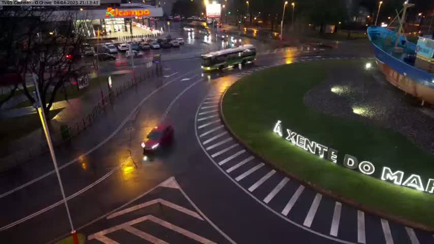 A wet roundabout at night shows a bus and several cars driving around a large, illuminated sign next to a boat sculpture.