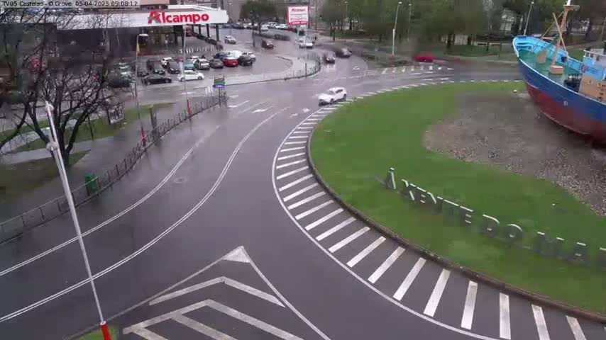 A wet roundabout with a large boat sculpture in the center, surrounded by a parking lot and roadways with several cars.