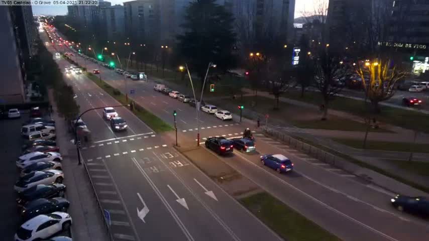 An aerial view of a city street at dusk shows several cars driving on a multi-lane road that intersects with a cross street, with parked cars lining the side of the road and a grassy median separating the lanes.