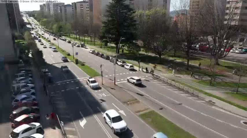 A sunny day view of a multi-lane road with cars driving in both directions, lined with buildings and trees, and pedestrians walking on a nearby sidewalk.