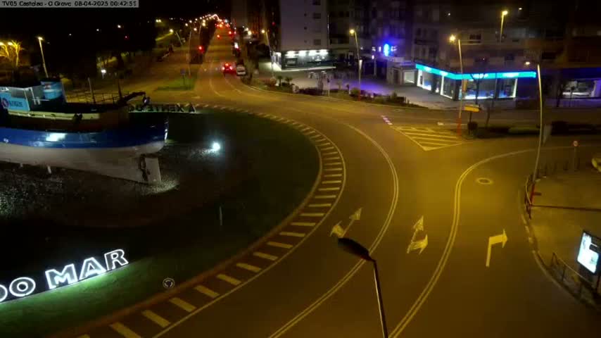 At night, a roundabout with a large model boat at its center is illuminated, surrounded by streets and buildings.