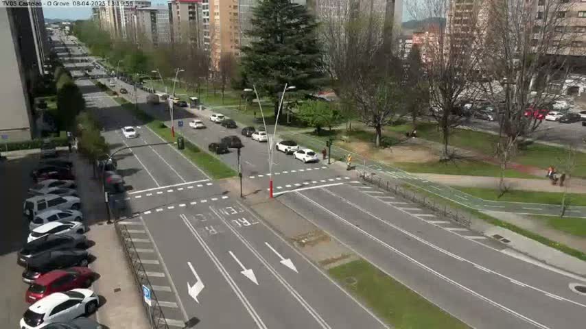 A sunny day shows a high-angle view of a multi-lane road intersection with cars, surrounded by buildings, trees, and a paved pedestrian/bicycle path.