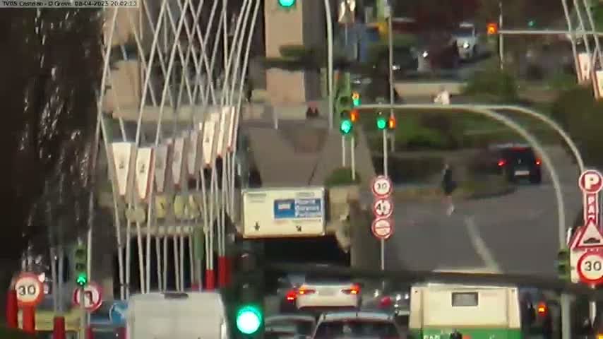A nighttime view of a busy city intersection with several cars and pedestrians, under partly cloudy skies.