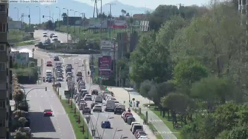 A sunny day shows a highway with multiple lanes of slow-moving traffic, curving around a roundabout surrounded by lush green trees and buildings.
