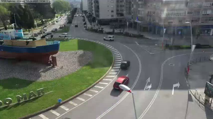 A large blue fishing boat sits on land beside a roundabout with several cars driving through it on a sunny day.