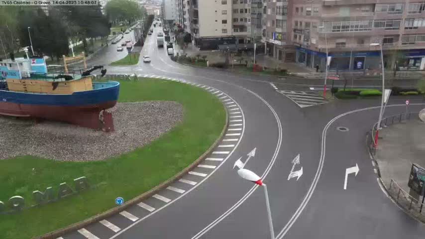 A large fishing boat sits on a grassy area next to a wet roundabout with several cars visible in the background.